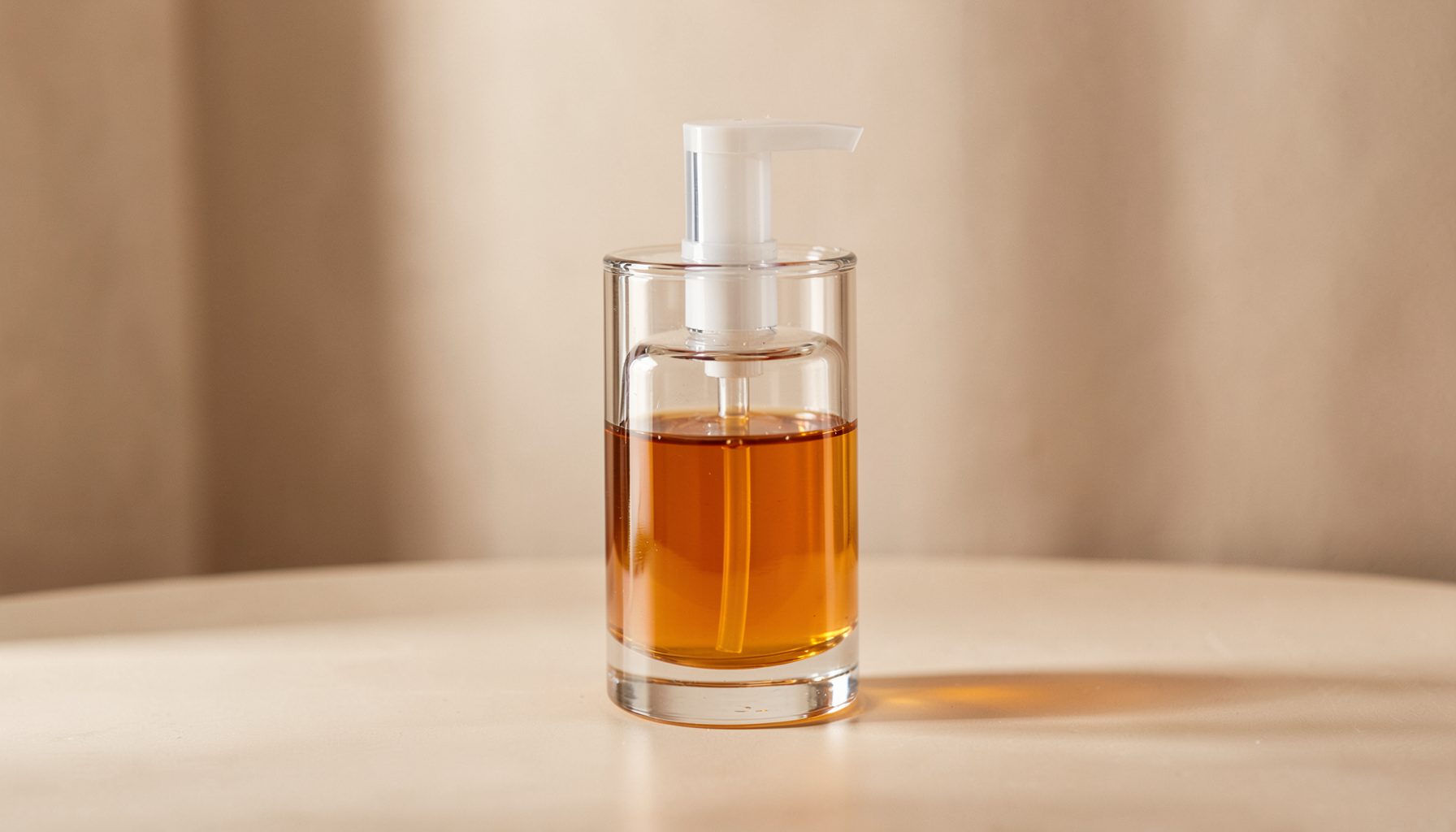 Clear glass dispenser with amber liquid soap, soft natural light, warm beige and brown tones, clean minimalist setting